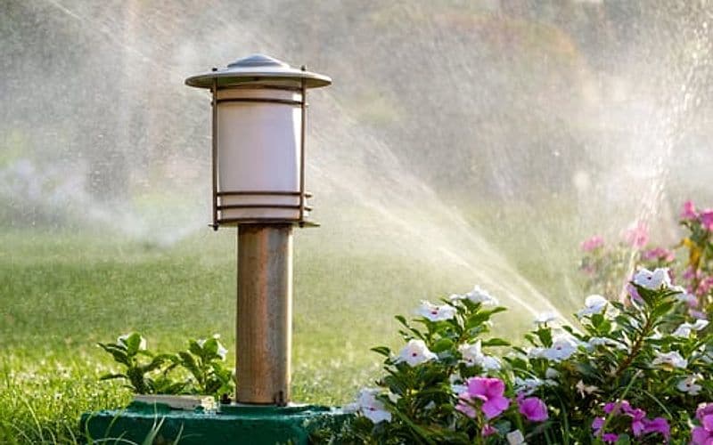 Irrigation sprinkler watering a lawn beside planted borders and a landscape light at dusk