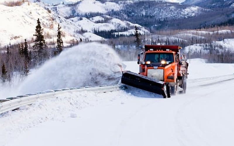 Snow plow clearing a road during a winter storm on a tree-lined route